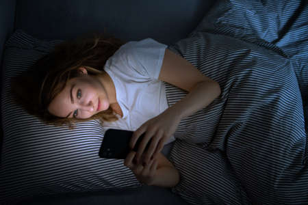 Young woman in bed holding a phone, tired and exhausted, blue light straining her eyes, messing up her circadian rhytmの写真素材