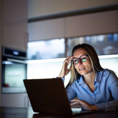 Pretty, middle-aged woman working late in the day on a laptop computer at home, running a business from home, working remotely - getting frustratedの写真素材