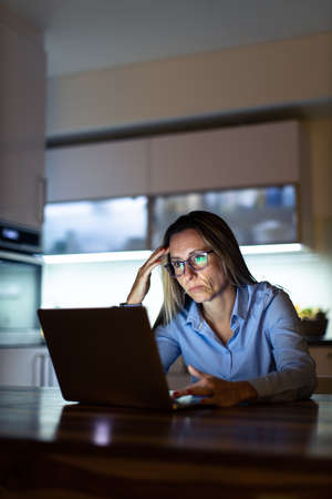 Pretty, middle-aged woman working late in the day on a laptop computer at home, running a business from home, working remotely - getting frustratedの写真素材