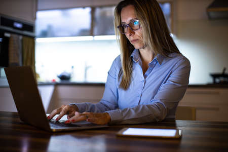 Pretty, middle-aged woman working late in the day on a laptop computer at home, running a business from home, working remotely - getting frustratedの写真素材