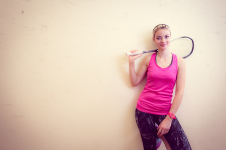 Cute young woman with a racket leaning against a wall in a squash court, ready for the gameの写真素材