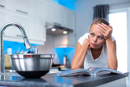 Pretty, young woman in her modern, clean and bright kitchen, thinking about what to cook for her family, looking quite desperate (color toned imagey; shallow DOF)の写真素材