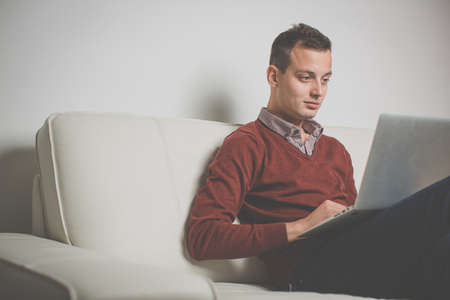 Sleepy young man sitting on a sofa and trying to get some work done on his laptop computer, yawningの写真素材
