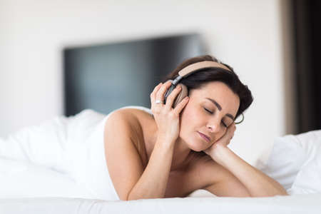 Pretty, young woman listening to music on her hi-fi headphones in bed, relaxingの写真素材