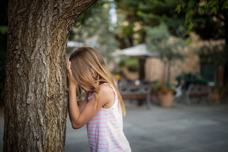 Little girl playing hide and seek with friends, counting by a tree, waiting for them to hideの写真素材