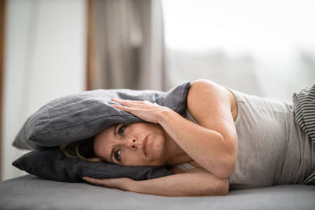 Sleepless lady covering ears with pillow. Noisy neighbors, tinnitus, insomnia or stress concept. Tired woman can't sleep. Awake in bed after coming home from her shift, work.の写真素材
