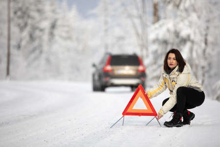 Pretty, young woman setting up a warning triangle and calling for assistance after her car broke down in the middle of nowhere on a freezing winter dayの写真素材