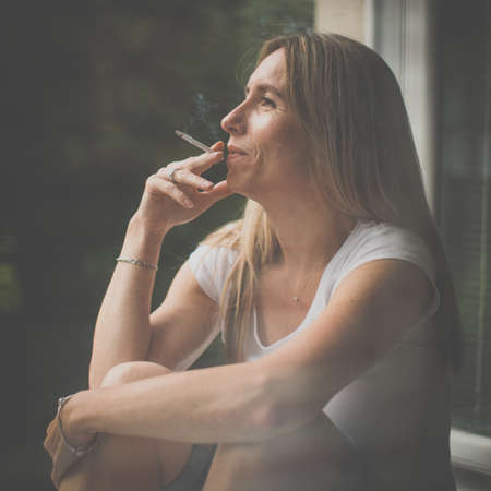 Mid-aged woman smoking a cigarette at home, getting her nicotine daily dose, unable to resist to her unhealthy habitの写真素材