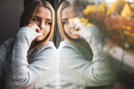 Depressed and anxious young woman sitting by a large window, feeling blue, sad, uncertainの写真素材
