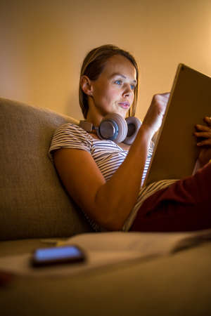 Pretty, young woman working at home in the evening - using her tablet computer as well as bluetooth headphones, writing down some ideas for tomorrowの写真素材