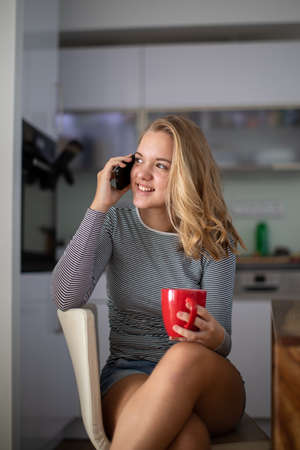 Teenage girl calling on her cell phone while having a cup of tea in modern kitchen setting (shalllow DOF, color toned image)の写真素材
