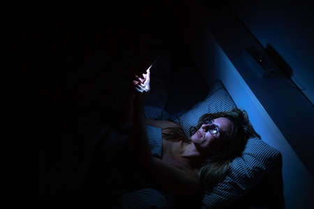 Pretty, middle-aged woman using her cell phone in bed at night - unhealthy blue light exposureの写真素材