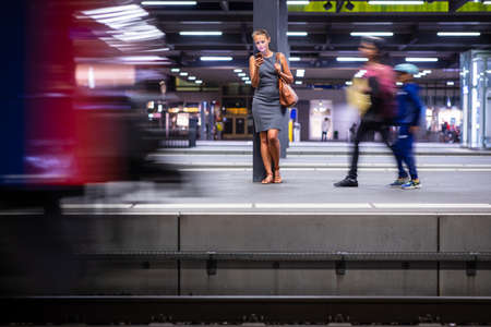 Pretty, young female commuter waiting for her daily train in a modern trainstation, using her cellphone while waiting (color toned image)の写真素材
