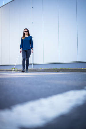 Young woman with impaired vision walking on city streets, using her white cane to navigate the urban space better and to get to her destination safelyの写真素材