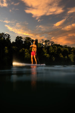 Handsome young man on a paddle board. Getting a great exercise on a lovely river in warm evening sunlightの写真素材
