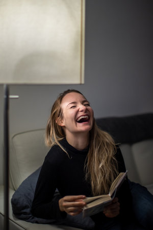 Avid reader laughing. Young attractive girl happy positive smile sit sofa home relax read book story novelty laugh humor jokeの写真素材