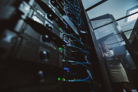 IT engineer standing amid working server racks doing routine maintenance check and diagnostics using laptop computer (color toned image)の写真素材