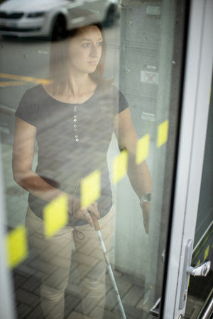 Young woman with impaired vision walking on city streets, using her white cane to navigate the urban space better and to get to her destination safelyの写真素材