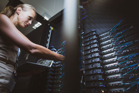 IT engineer standing amid working server racks doing routine maintenance check and diagnostics using laptop computer (color toned image)の写真素材
