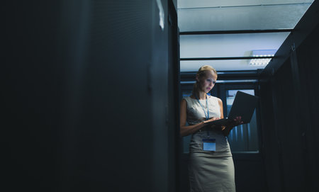IT engineer standing amid working server racks doing routine maintenance check and diagnostics using laptop computer (color toned image)の写真素材