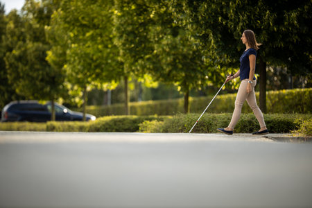 Young woman with impaired vision walking on city streets, using her white cane to navigate the urban space better and to get to her destination safelyの写真素材