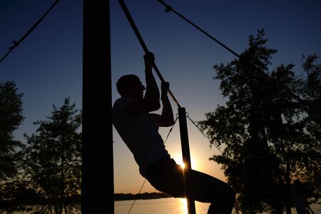 young man doing pull ups on horizontal bar outdoors during sunset - fitness, sport, exercising,...