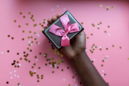 African woman's hand taking a craft textured gift box with ribbon bow surrounded by stars and snowflakes on a coral background, from above..の写真素材