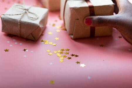African woman's hand taking a craft textured gift box with ribbon bow surrounded by stars and snowflakes on a coral background, from above..の写真素材