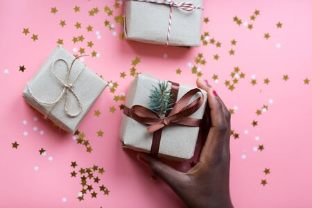 African womans hand taking a craft textured gift box with ribbon bow surrounded by stars and snowflakes on a coral background, from aboveの写真素材