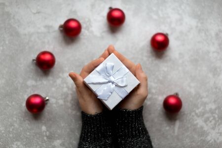 Woman's hand is taking a craft textured gift box with ribbon bow by unfocused vinous Christmas balls on a gray background, from above..の写真素材