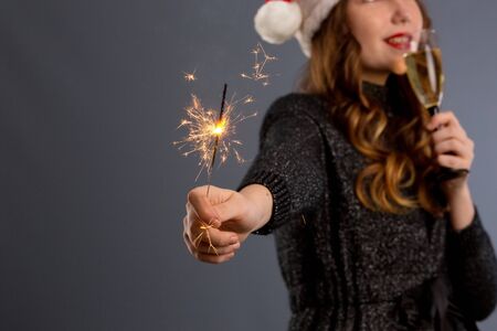 Attractive cheerful girl with curly hair in Christmas hat smiling, holding sparkler and drinking champagne, isolated on gray background..の写真素材