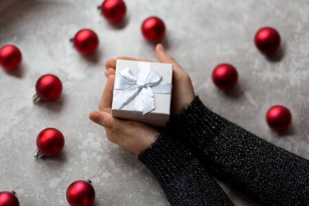 Woman's hands are holding a craft textured gift box with ribbon bow by unfocused vinous Christmas balls on a gray background, from above..の写真素材