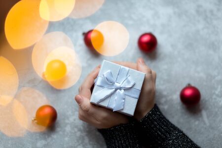 Woman's hands are holding a craft textured gift box with ribbon bow by unfocused vinous Christmas balls on a gray background, from above..の写真素材
