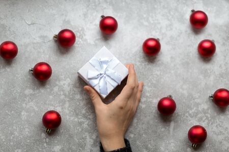 Woman's hand is taking a craft textured gift box with ribbon bow by unfocused vinous Christmas balls on a gray background, from above..の写真素材