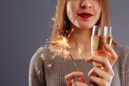 Attractive cheerful lovely girl with long light hair smiling, holding Bengal light and drinking champagne, isolated on gray background..の写真素材