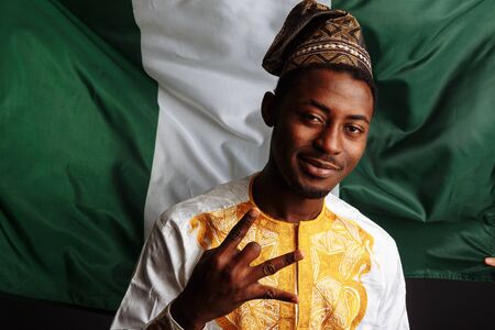 African man with traditional clothes is posing before the camera, smiling and has fun before the flag of Nigeriaの写真素材