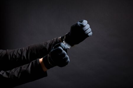 closeup of the hands of a burglar in black gloves with handcuffs on a black background.の写真素材