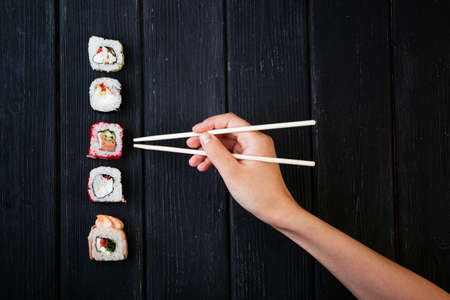 Female hand takes chopsticks sushi rolls with Chinese chopsticks. Lying on a black wooden board. View from above.の写真素材
