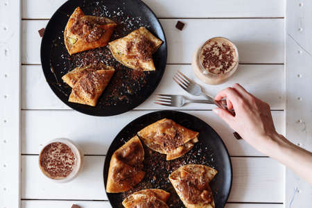 Woman's hand decorates the table with pancakes with chocolate, kiwi and banana. Black plate, white table. From above.の写真素材