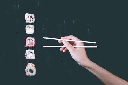 Female hand takes chopsticks sushi rolls with Chinese chopsticks. Lying on a black wooden board. View from aboveの写真素材