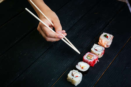 Female hand takes sushi rolls with Chinese chopsticks. Lying on a black wooden board. Lying on a...