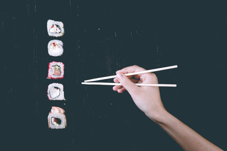 Female hand takes chopsticks sushi rolls with Chinese chopsticks. Lying on a black wooden board. View from aboveの写真素材