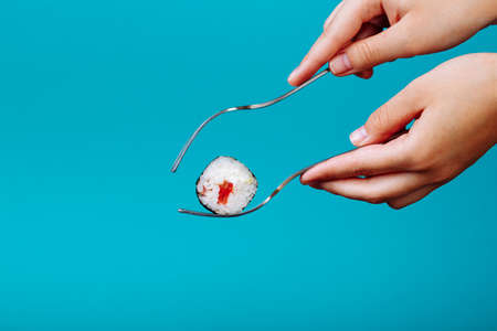 Woman hands holding fresh sushi roll with a fork, isolated on blue background. How to eat sushi without chopsticksの写真素材
