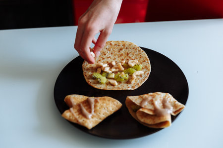 Womans hand decorates pancakes with a fruits kiwi and banana. Black plate, white tableの写真素材