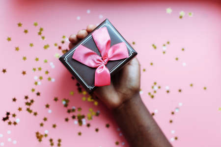 Hand of African woman holding present box with a bow on pastel pink background with stars and snowflakes confetti. Selective focusの写真素材