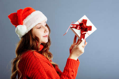 Beautiful lady in Santa hat is holding presents, looking at camera and smiling, isolated on gray backgroundの写真素材