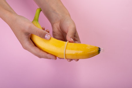 Female hand put on condom to banana on pink background. The concept of contraception.の写真素材