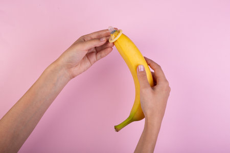 Female hand put on condom to banana on pink background. The concept of contraception.の写真素材