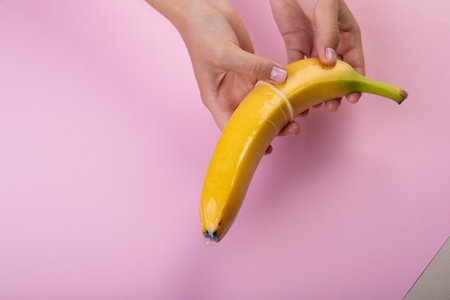 Female hand put on condom to banana on pink background. The concept of contraception.の写真素材