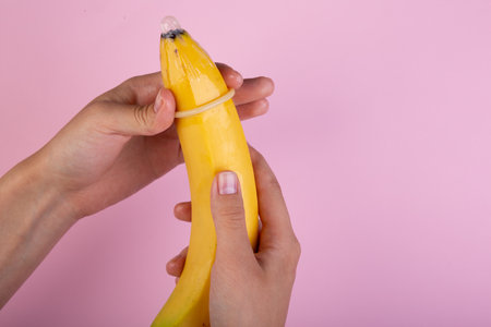 Female hand put on condom to banana on pink background. The concept of contraception.の写真素材
